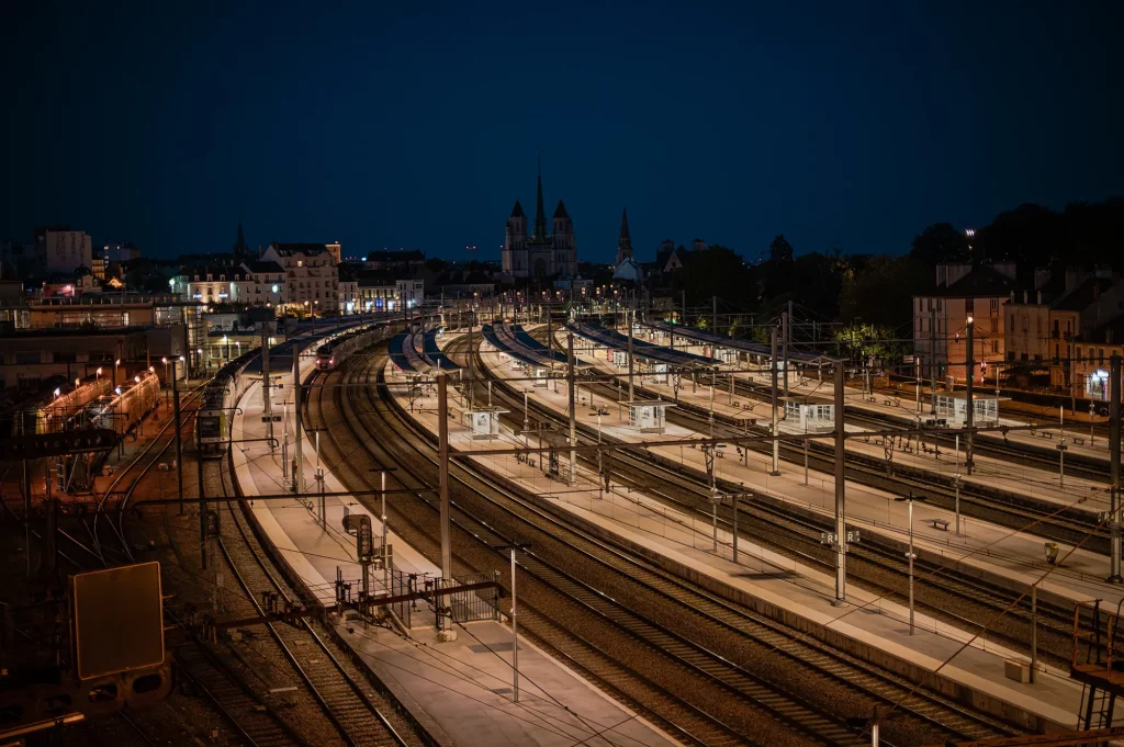 Gare de Dijon éclairée la nuit, avec les voies ferrées et les clochers de la ville en arrière-plan